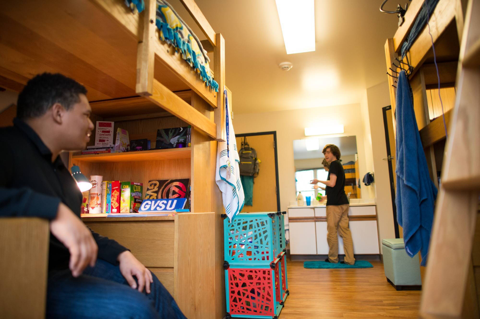 A cozy college Suite style bedroom features two lofted beds. A person sits on the lower bed, while another stands by a sink. The room has a warm, friendly atmosphere.